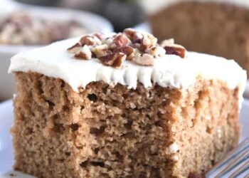 Square of frosted applesauce spice cake on white plate with pecans on top.