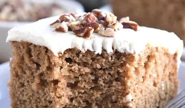 Square of frosted applesauce spice cake on white plate with pecans on top.