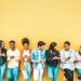 A group of people leaning against a yellow wall, holding and looking at their cell phones.