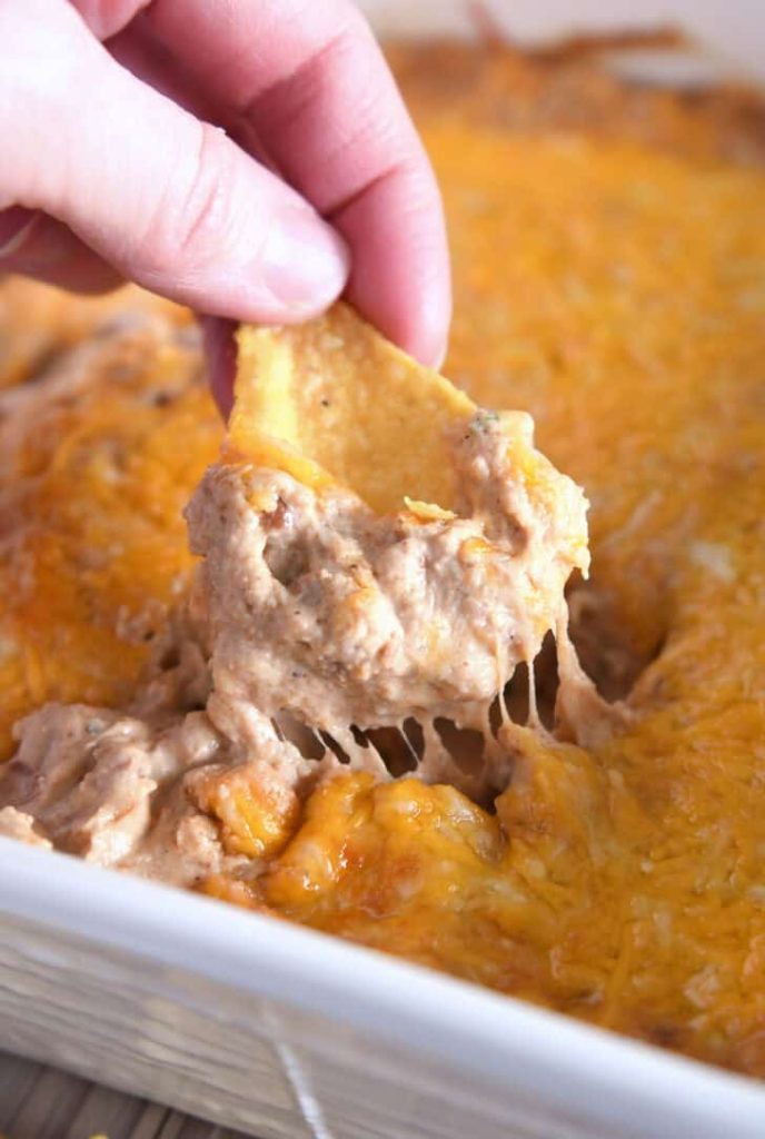 Fingers grabbing tortilla chip and dipping in cheesy, warm bean dip.