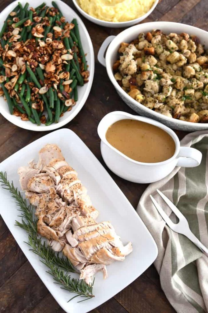 Top down view of platters of turkey, rosemary, gravy, stuffing, green beans and mashed potatoes.